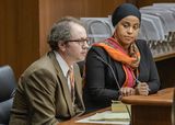 Mark Erpelding sits at the testifiers' table and looks ahead at someone out of view. Representative Hassan sits next to him and looks in his direction.